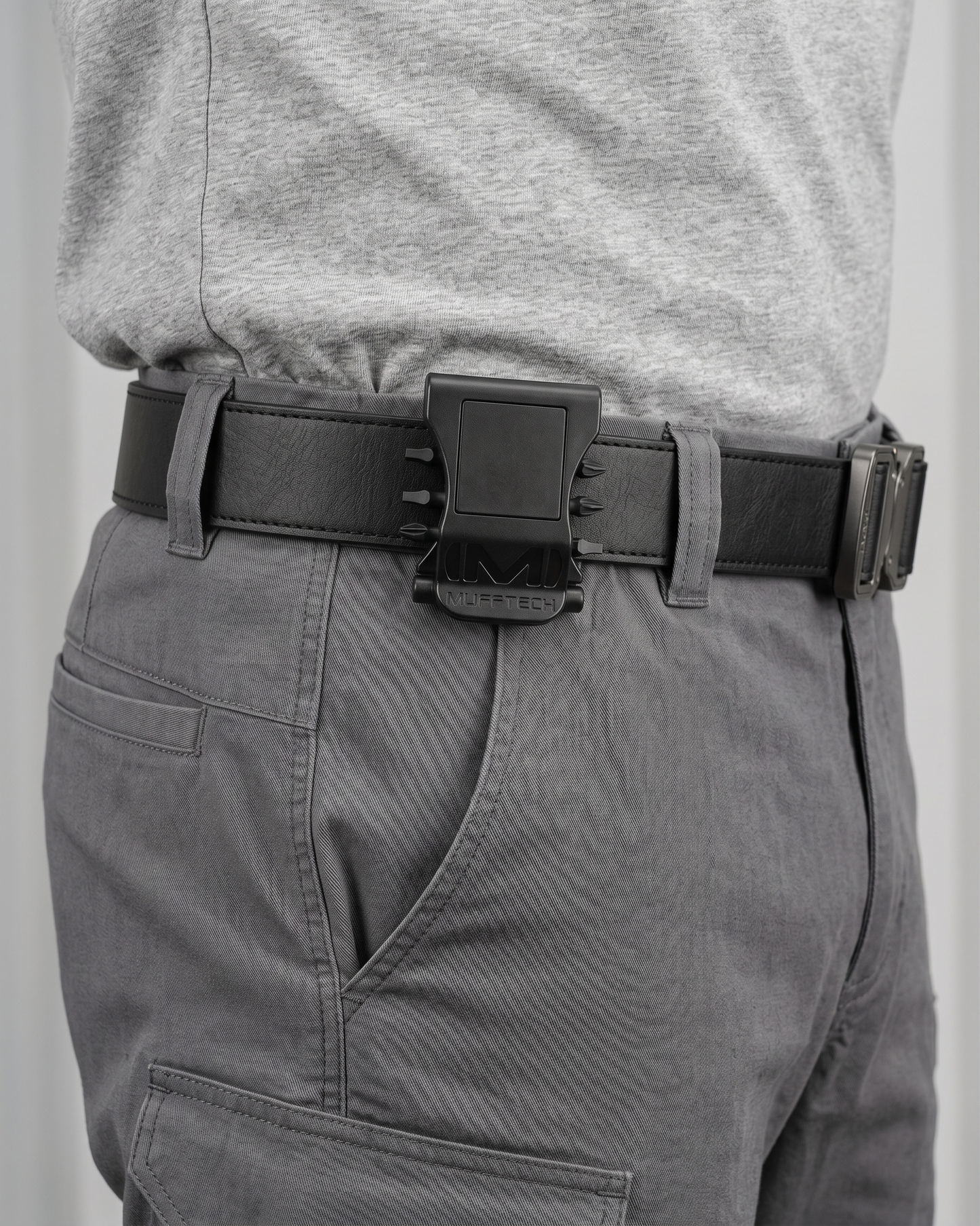 Person wearing a black belt with a large buckle on gray pants against a plain background